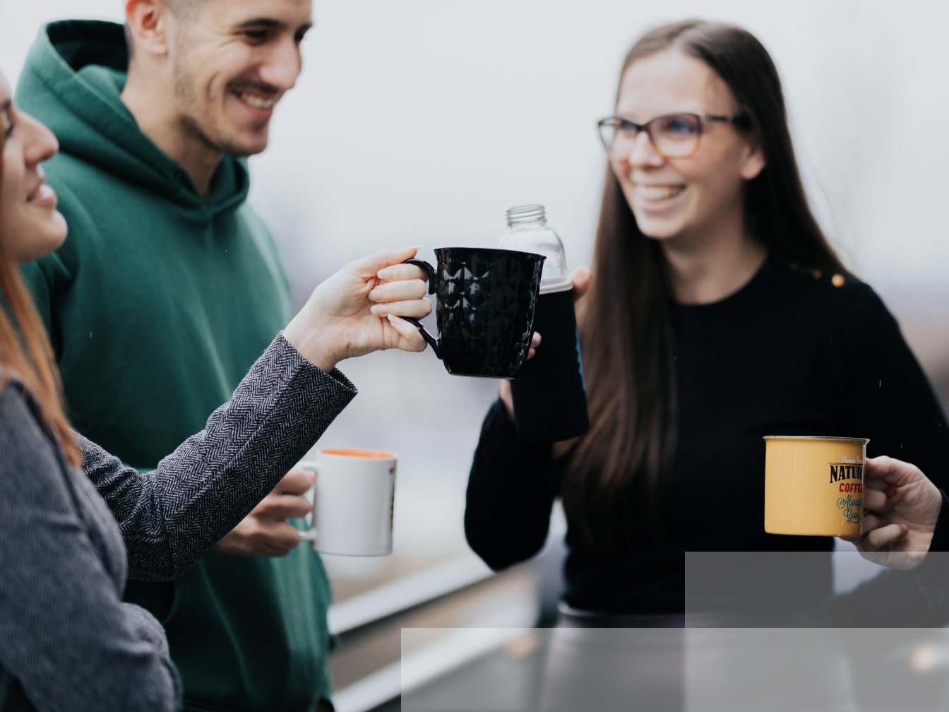 Four smiling people drinking coffee on a balcony, two of them toasting with a bottle of water and cups of coffee.
