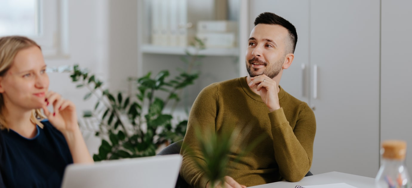 A man and a woman sitting in a meeting, with a plant in the background, out of focus.